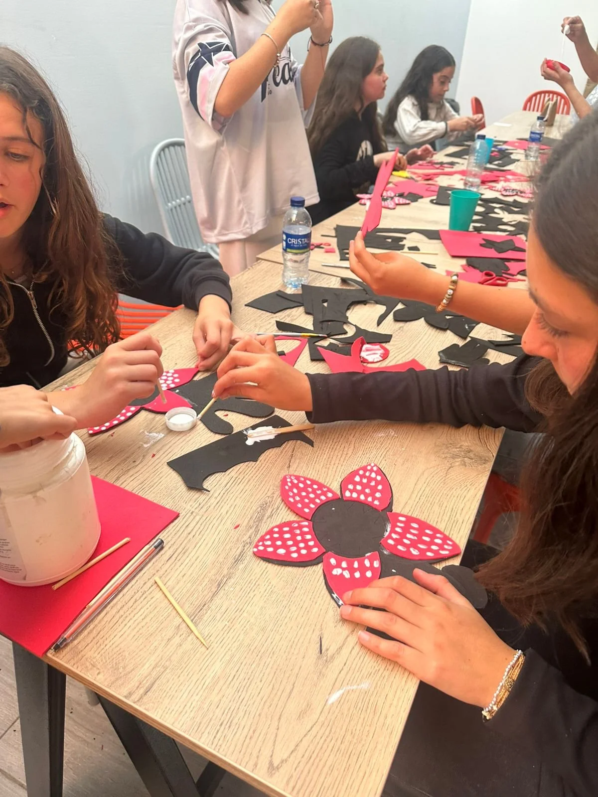 Niñas y mujeres decorando flores de papel con colores negro y rojo, en una mesa de manualidades, en un ambiente de sala o aula.