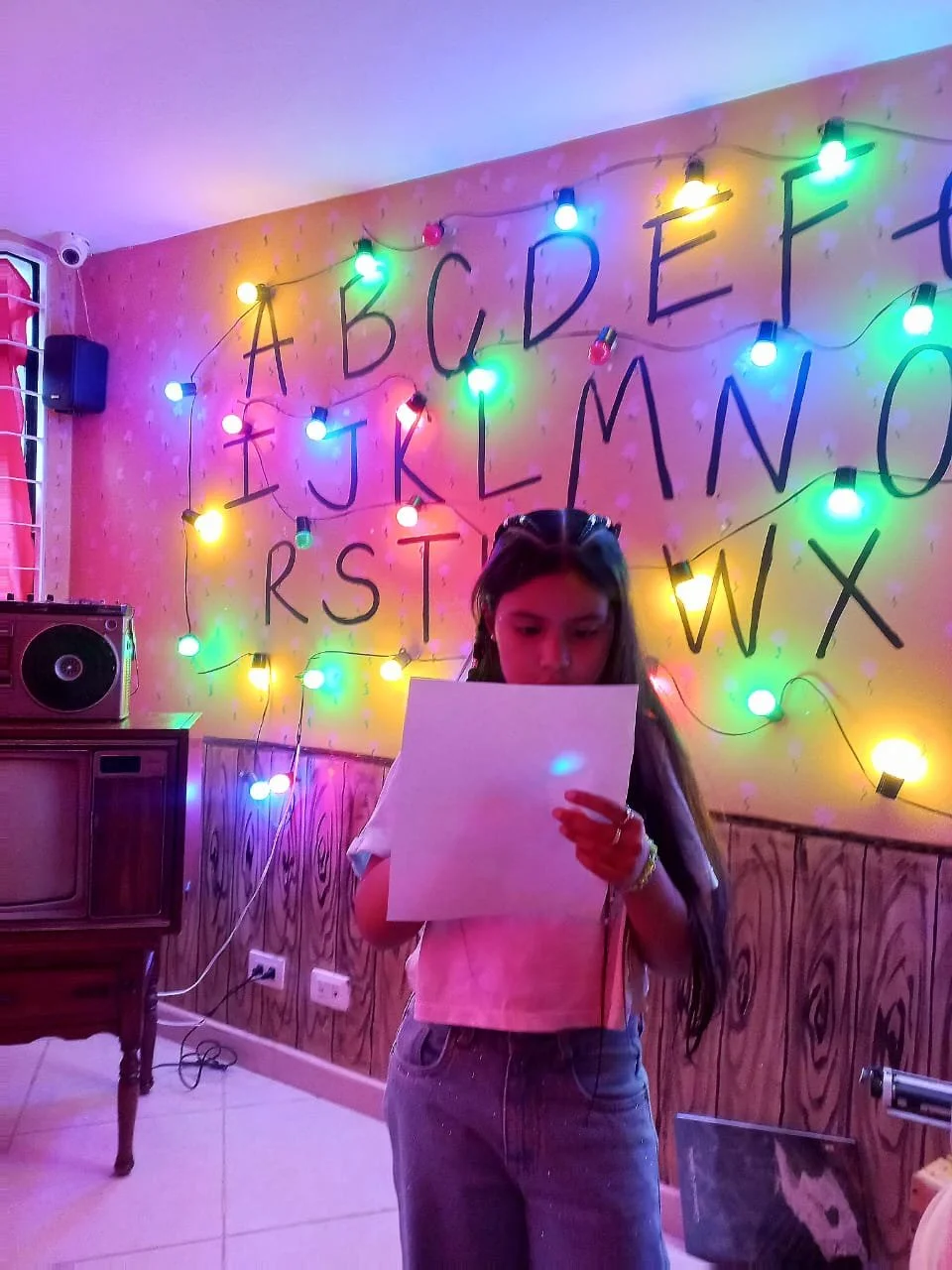 Joven leyendo un papel en una habitación decorada con luces de colores y letras del alfabeto en la pared