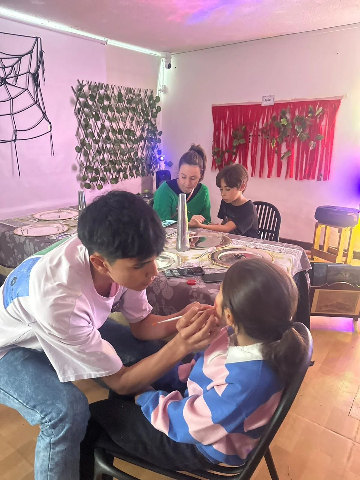 Un joven le hace un maquillaje a una niña mientras otras personas observan en una sala decorada con tela y plantas.