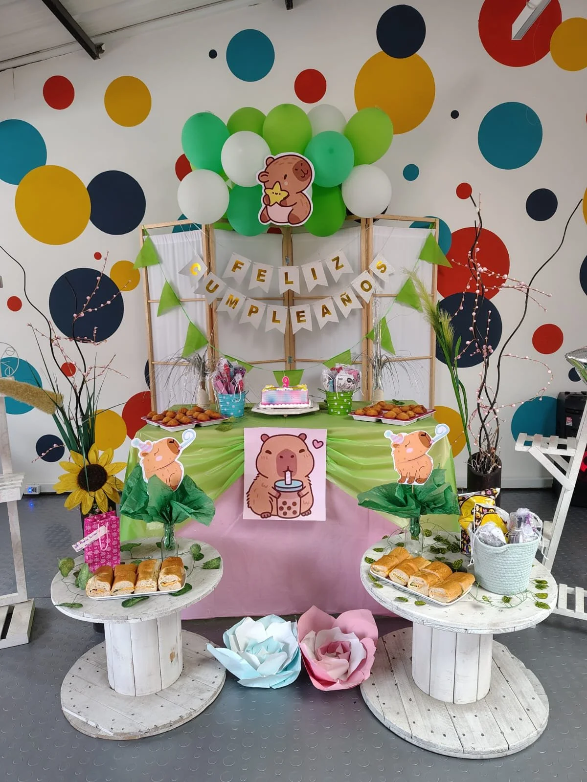 Decoración para fiesta de cumpleaños con temática de cerditos. Incluye globos verdes, blancos y otros colores, carteles de felicitaciones, figuras de cerditos y comida como panes enrollados y galletas. Hay globos de papel en forma de flores y ramas decorativas.