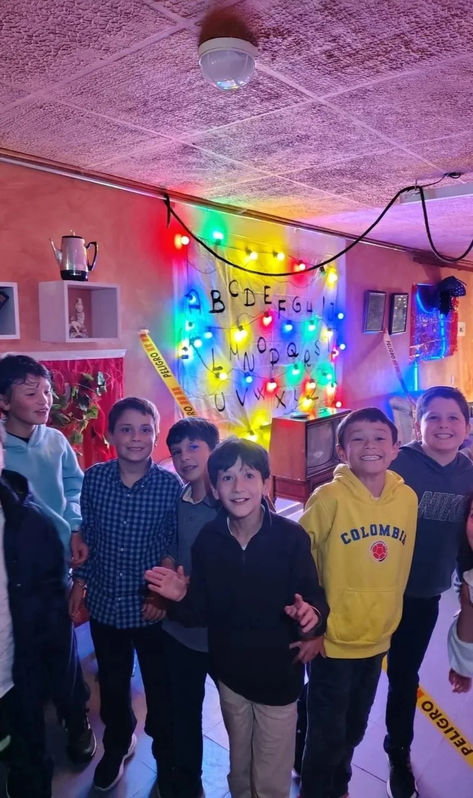 Niños sonriendo en una fiesta con decoraciones de luces, el tablero del Demogorgon de la serie Stranger Things y una señal de peligro en el fondo.