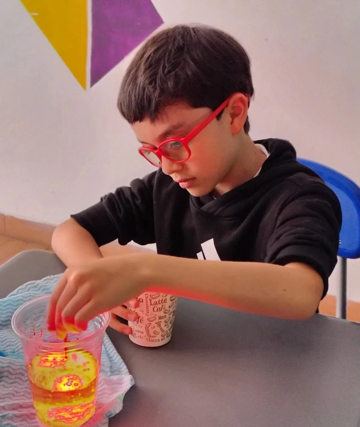 Un niño con gafas rojas colocando una pajilla en un vaso con bebida amarilla y hielo, sentado en una mesa en un ambiente interior.