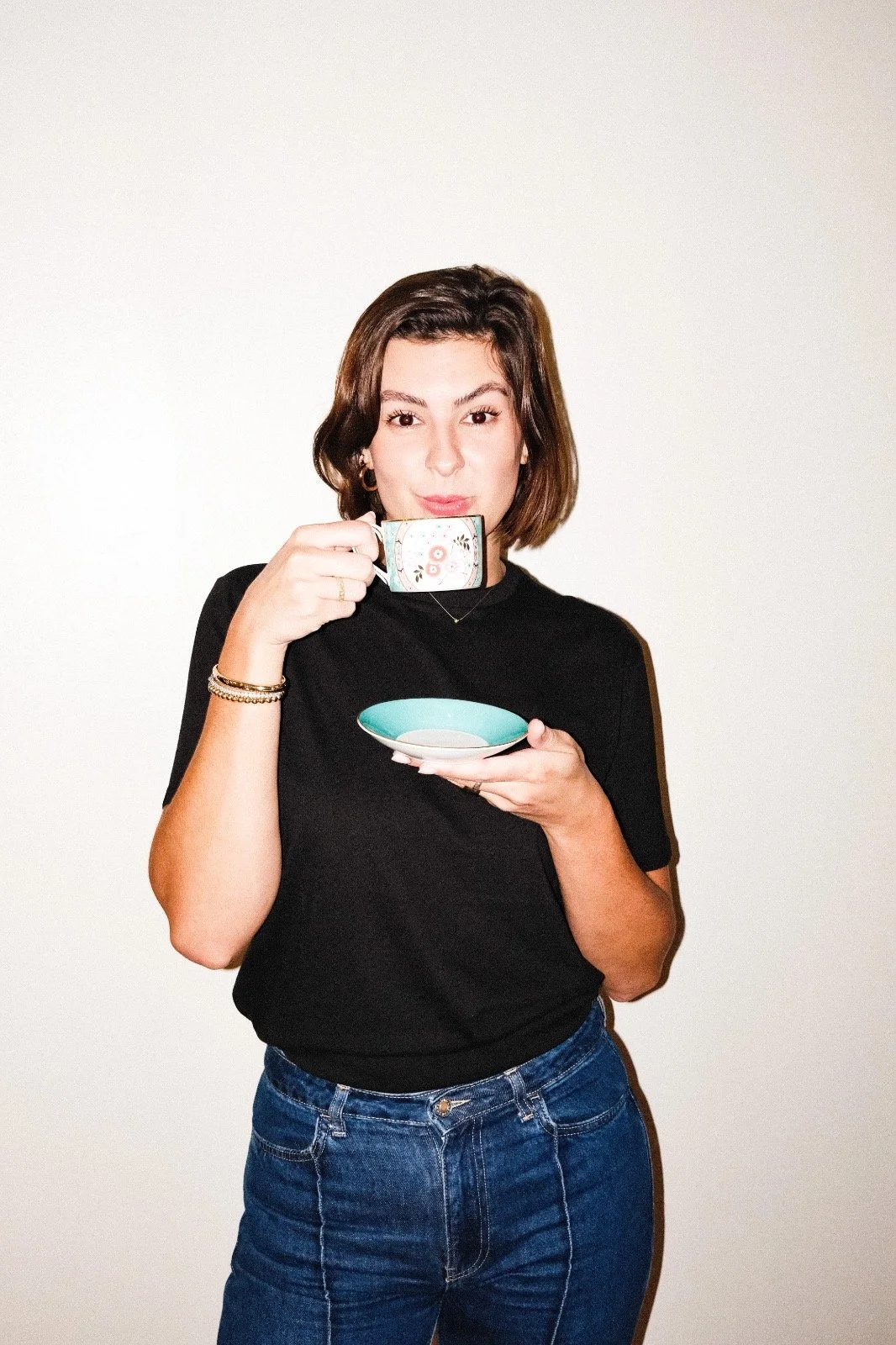 A woman with short dark hair wearing a black shirt and blue jeans, holding a tea cup in one hand and a saucer in the other, against a plain white background.