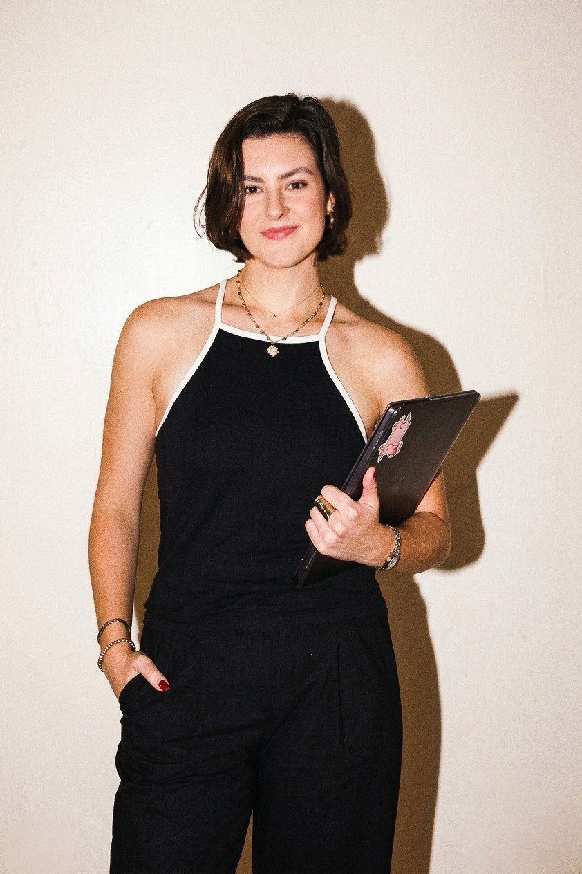 A woman with shoulder-length brown hair, wearing a sleeveless black top with white trim, holding a black laptop with a pink sticker, standing against a plain light-colored wall.