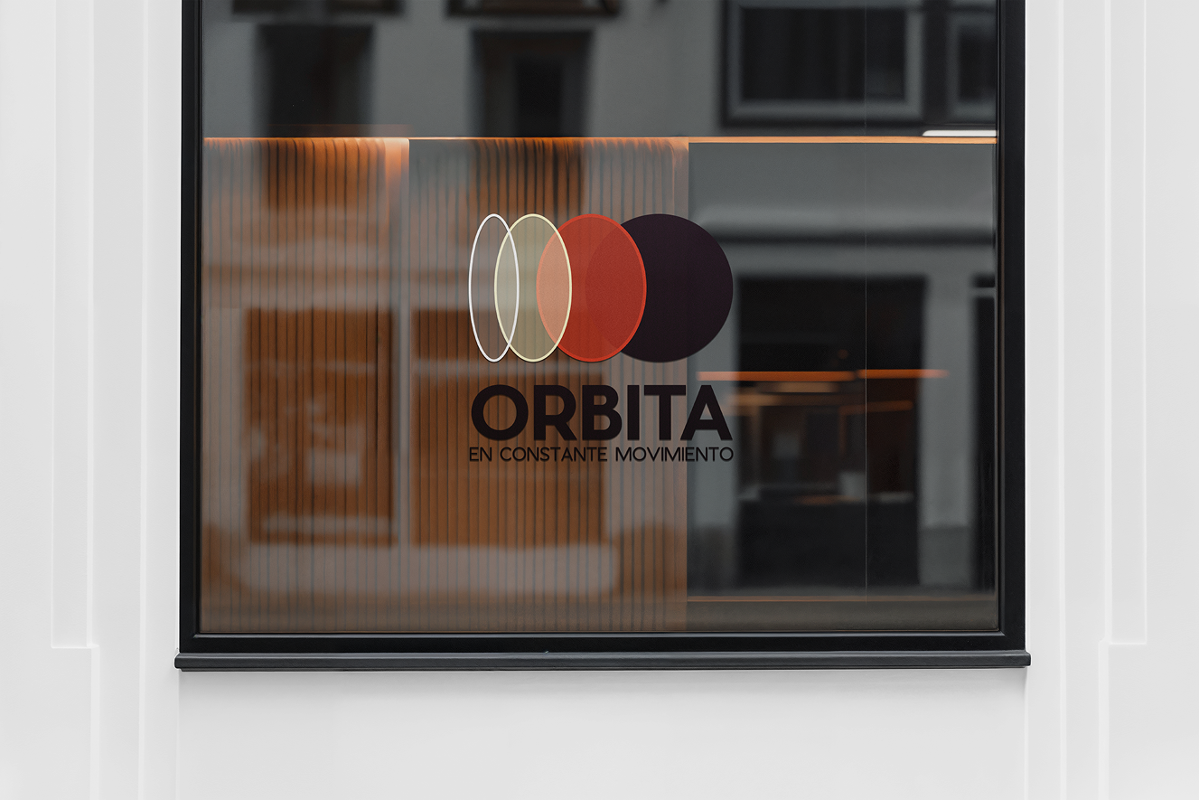 Glass door with a logo and text. The logo consists of five overlapping circles in white, cream, red, and black. Below the logo, the text reads "ORBITA" and "EN CONSTANCIA MOVIMIENTO." The background shows an interior with framed pictures on the wall.