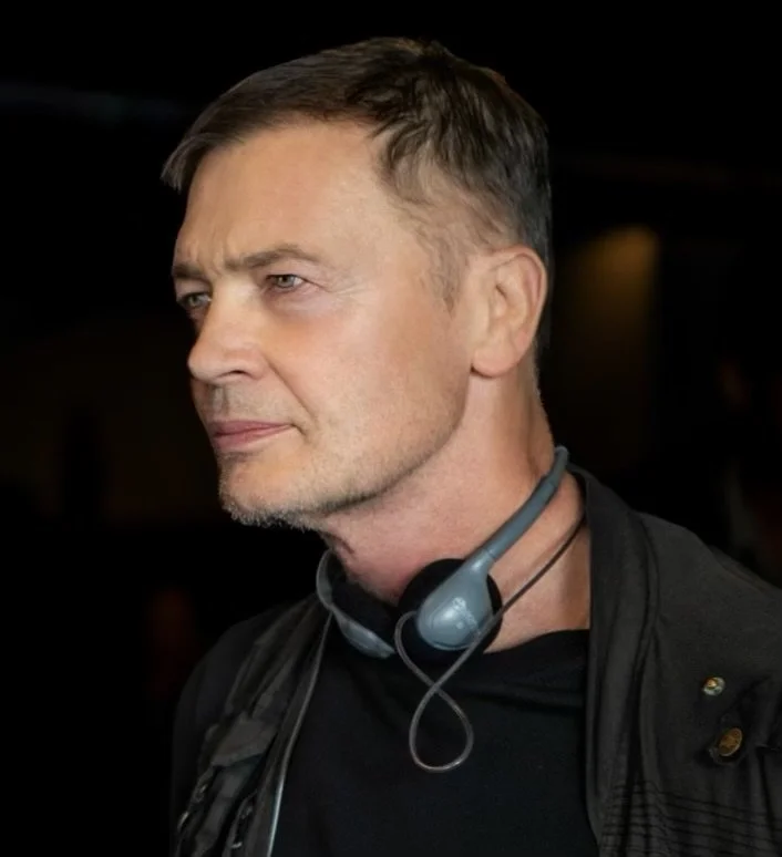 A middle-aged man with short brown hair, wearing a black jacket and a headset around his neck, looking to the left in a dark environment.