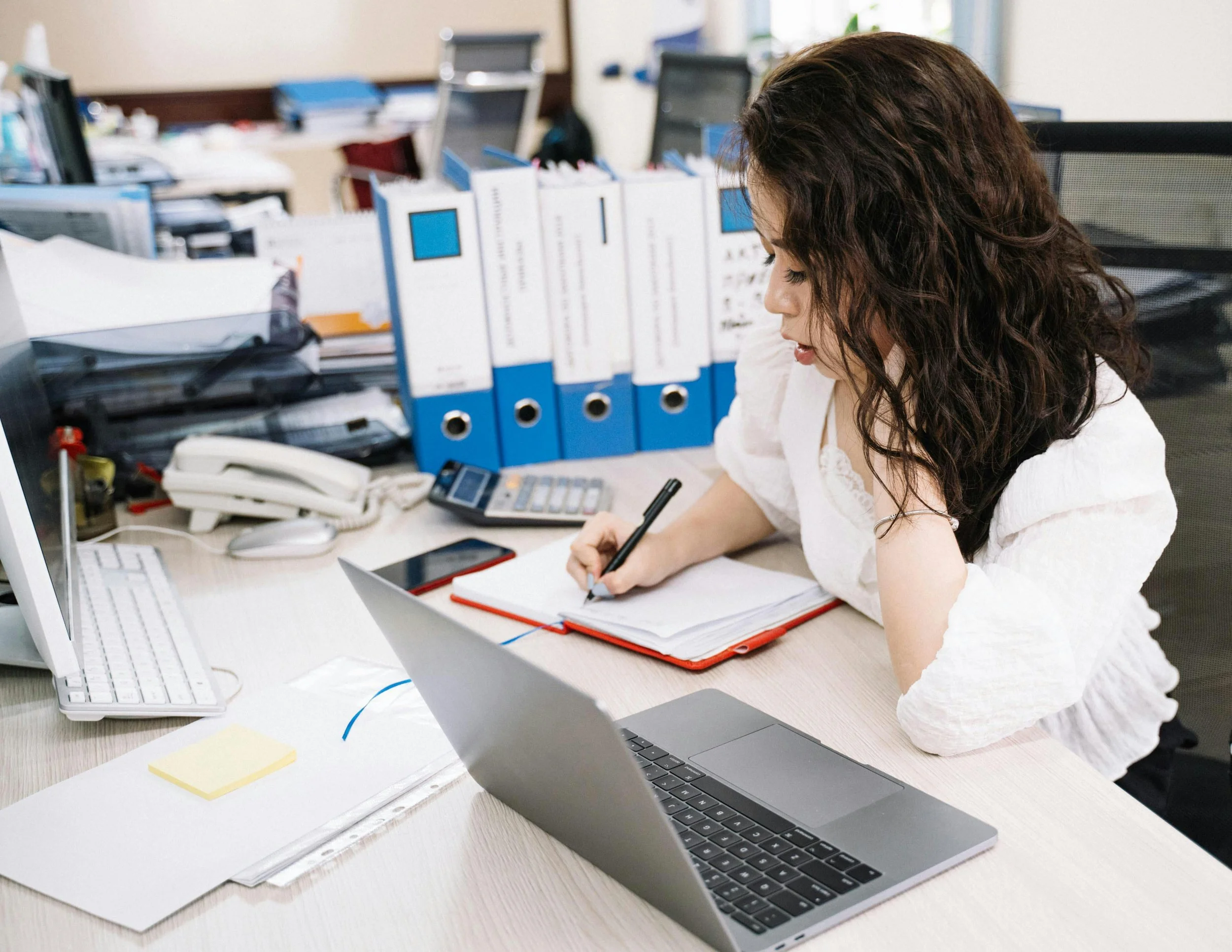 A woman with long, curly brown hair sitting at an office desk using a laptop and writing on a notepad. The desk has a computer monitor, a landline phone, a calculator, and stacked blue binders. The background shows more office desks with papers and supplies.