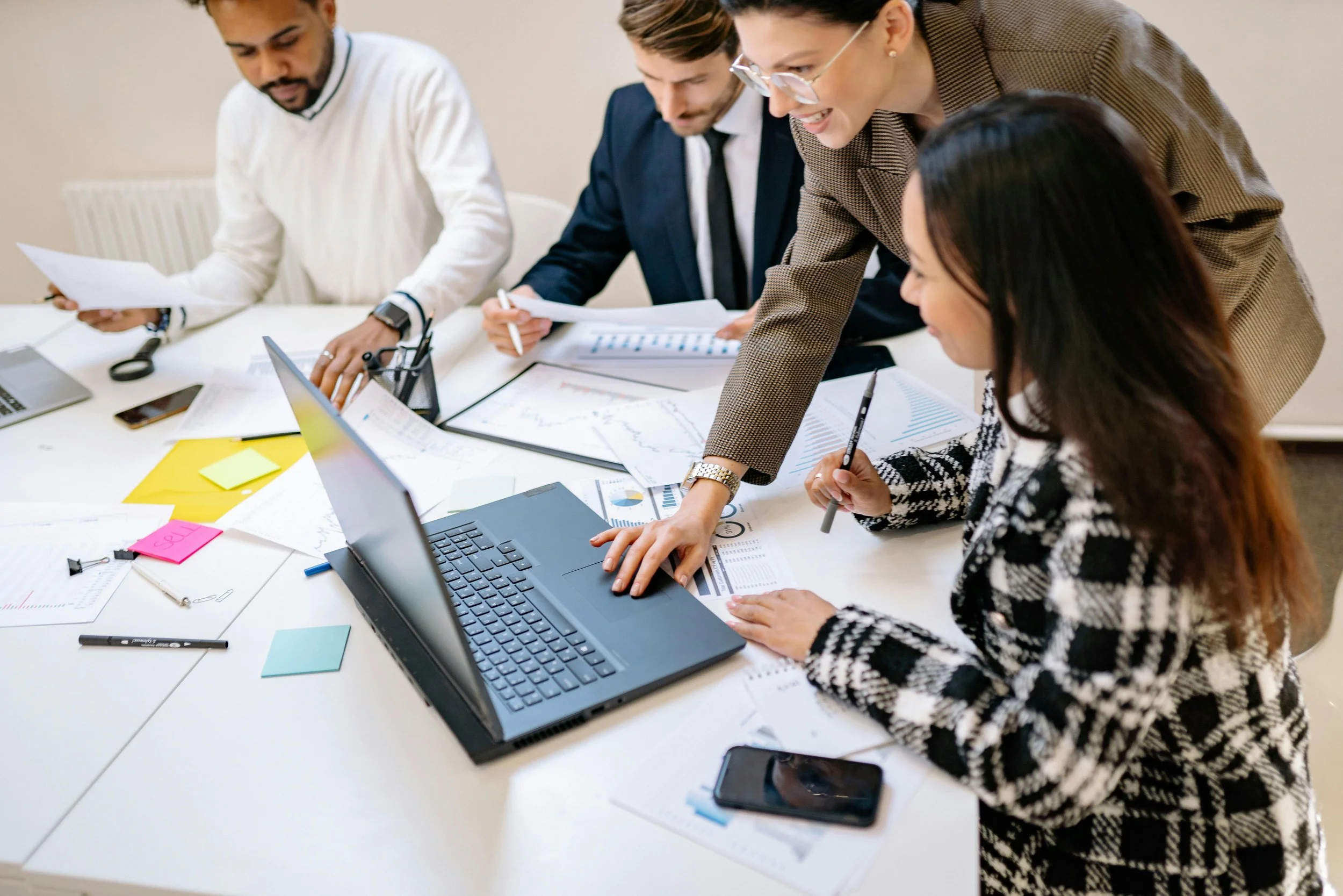 A group of five professionals in a meeting, discussing documents and data on a laptop and papers on the table, with sticky notes and writing utensils.