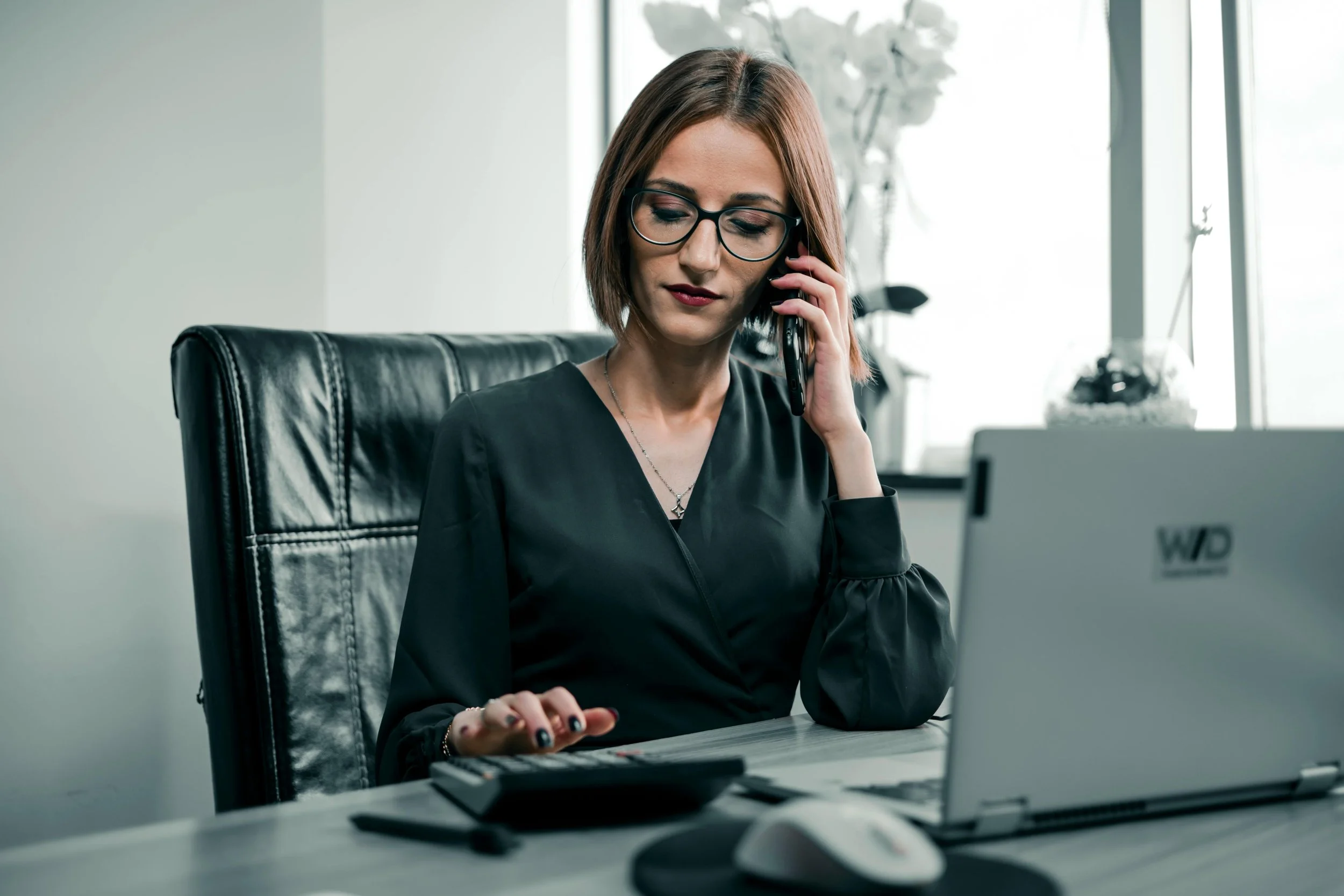 A woman with glasses talking on the phone in an office environment, seated at a desk with a computer and various office supplies.