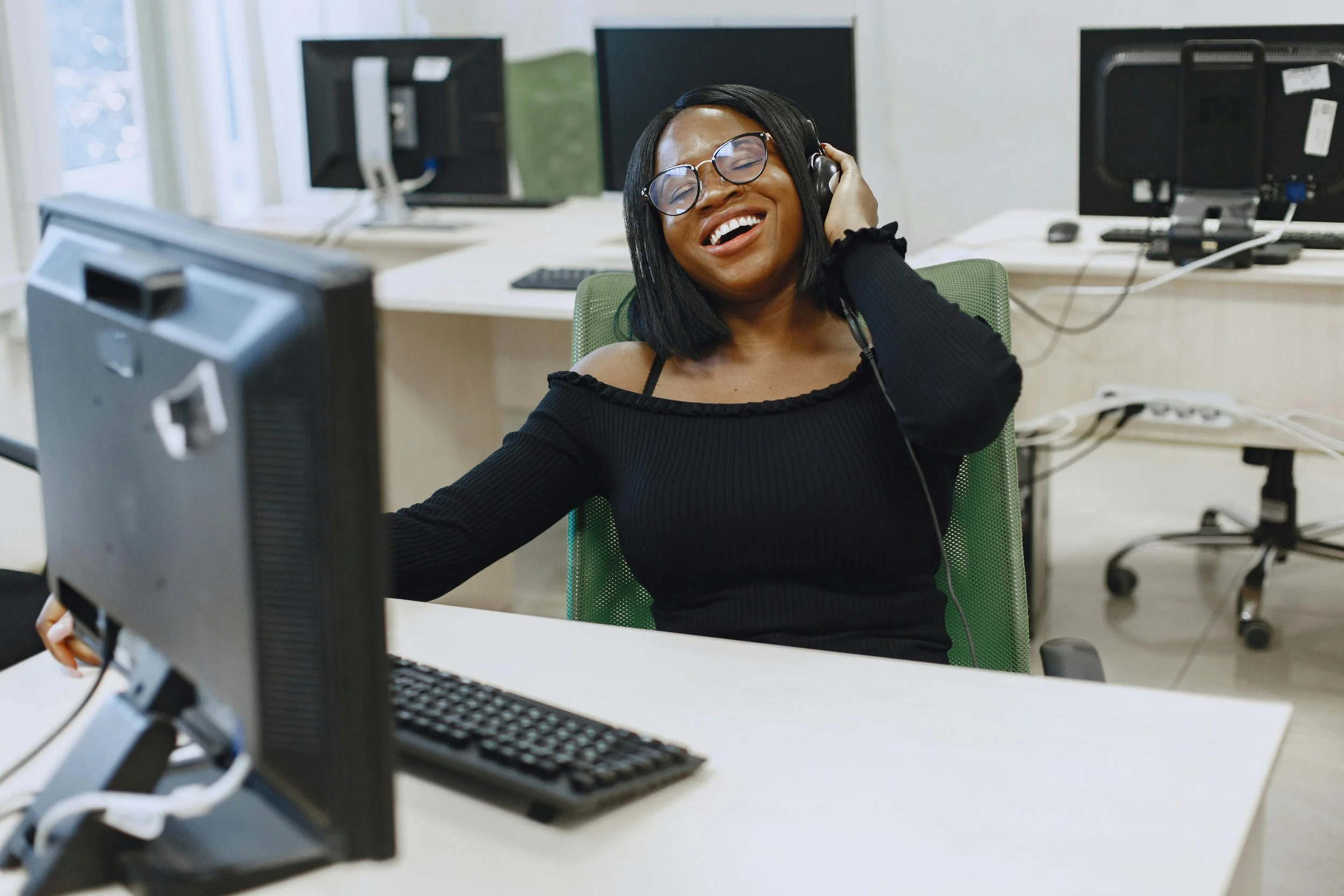 A woman with glasses and headphones smiling while sitting at a desk in an office with multiple computer monitors.