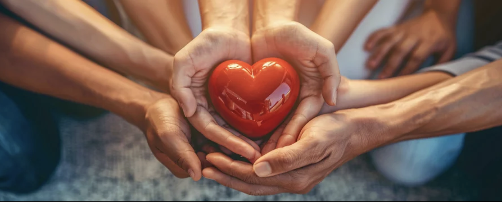 Multiple hands of different people gently holding a red, shiny heart-shaped object.