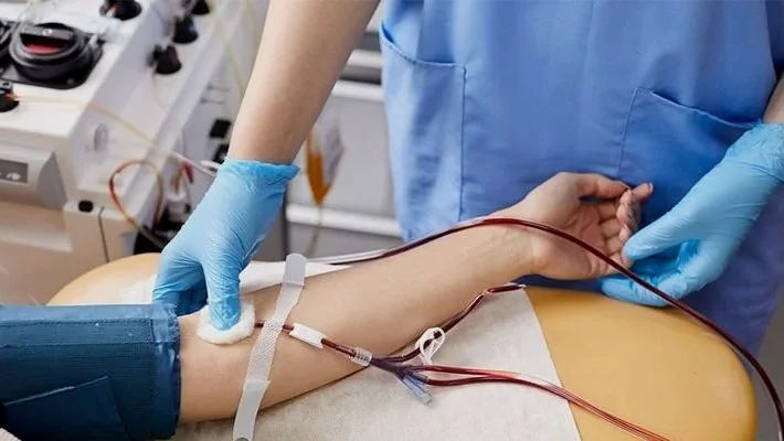 A healthcare worker in blue scrubs and gloves is drawing blood from a patient's arm, which is secured with a tourniquet and connected to medical tubes in a clinical setting.