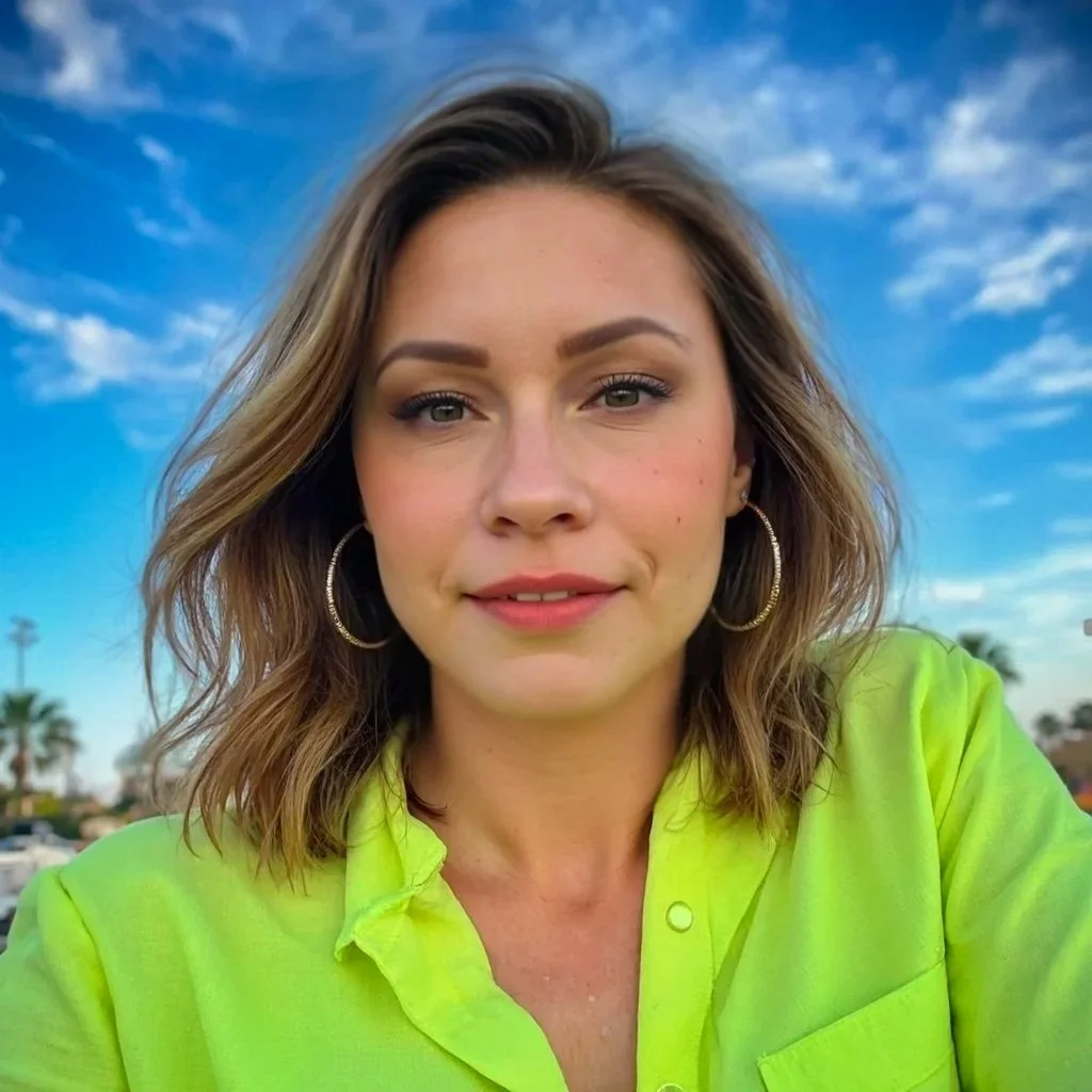 A woman with shoulder-length wavy hair taking a selfie outdoors against a blue sky with some clouds, wearing a bright yellow shirt and large hoop earrings.