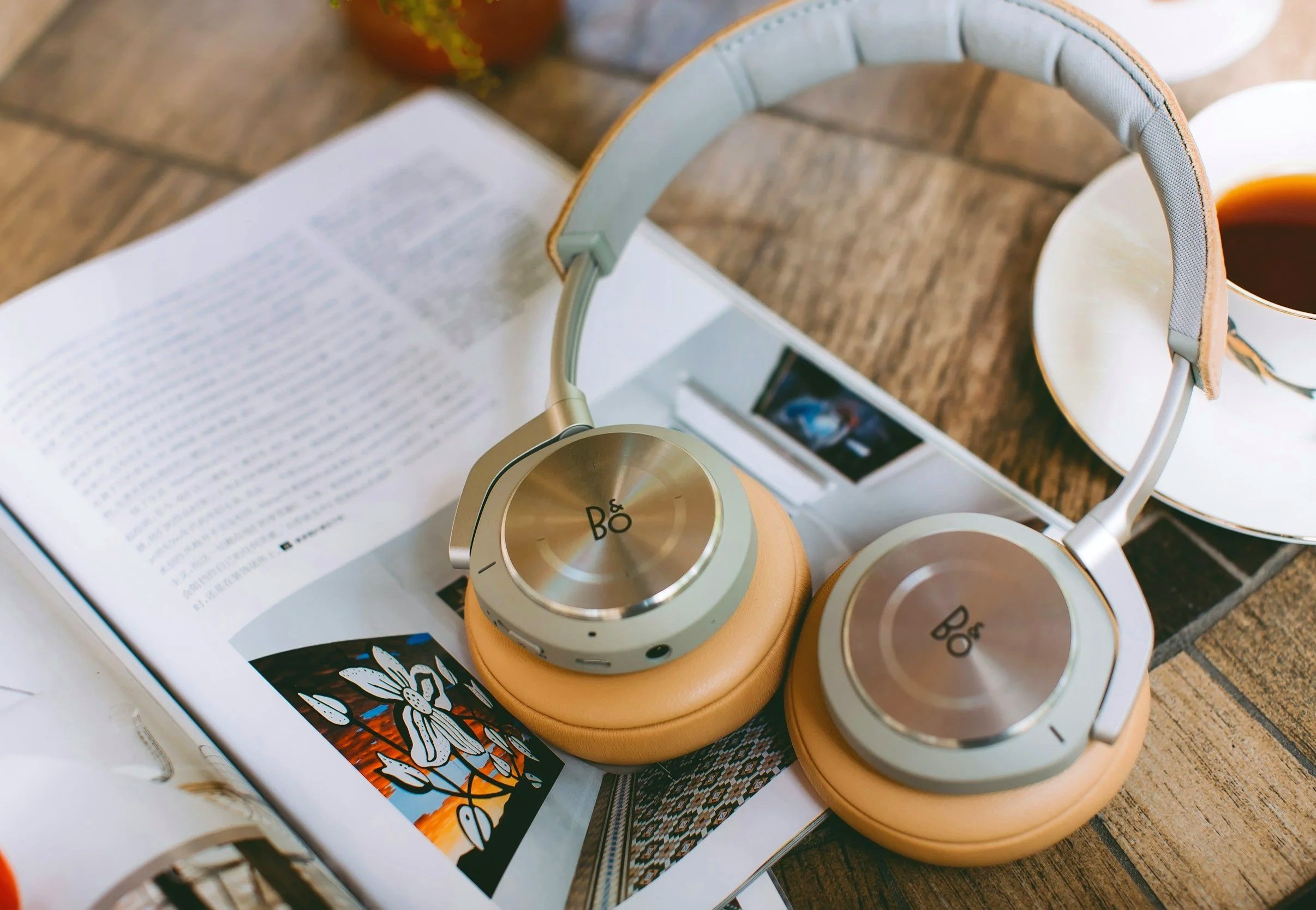 A pair of beige over-ear headphones with gold accents resting on a magazine or book on a wooden table, with a cup of coffee and a tumbler in the background.