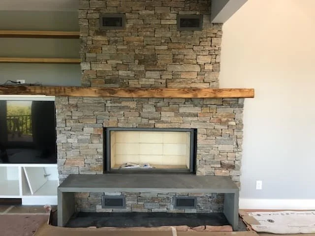 A modern living room fireplace with a stone surround, a wooden mantel, and a concrete hearth, with built-in speakers and a TV to the side.