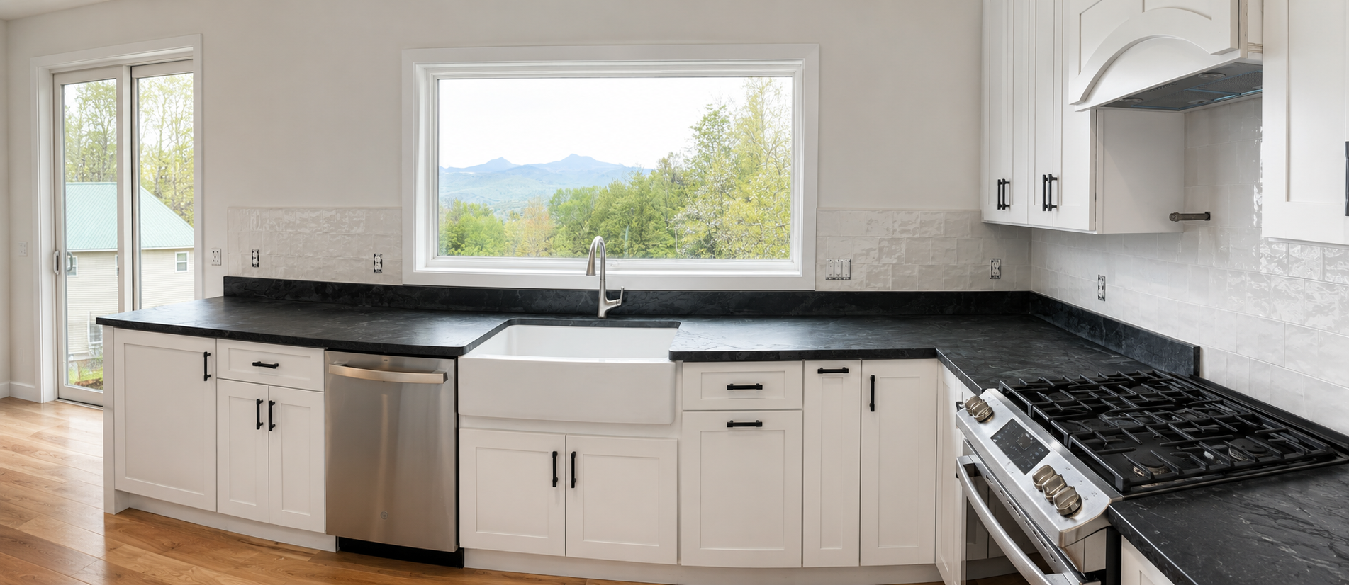 Kitchen with white cabinets, black countertops, a stainless steel dishwasher, a large window with a view of trees and mountains, a black stovetop, and hardwood floors.