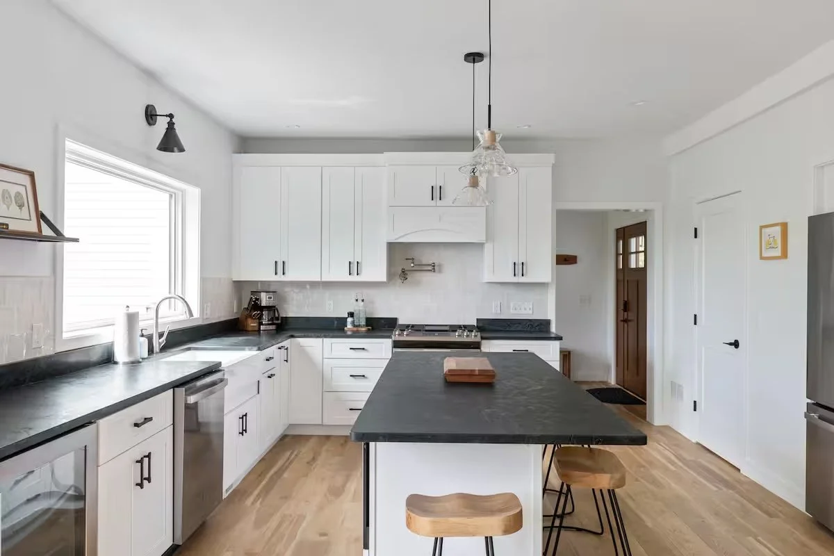 Modern white kitchen with black countertops and wooden stools, featuring a window over the sink, stainless steel appliances, and a central island.