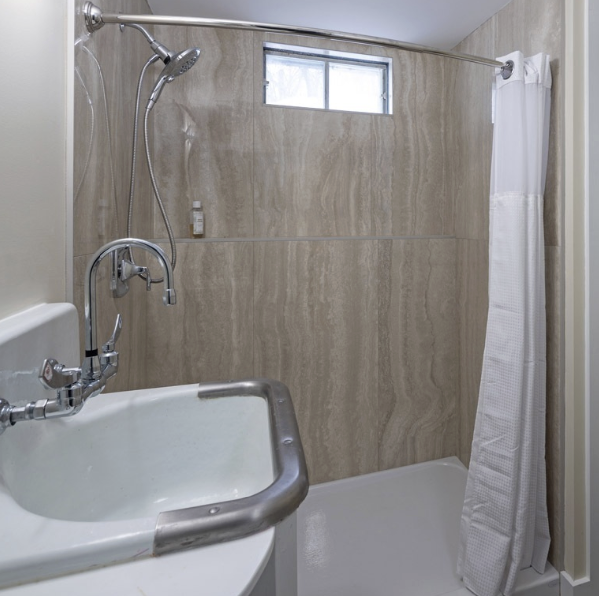 Small bathroom with a shower area, beige tiled walls, a small horizontal window, a white shower curtain, and a white sink with a faucet.