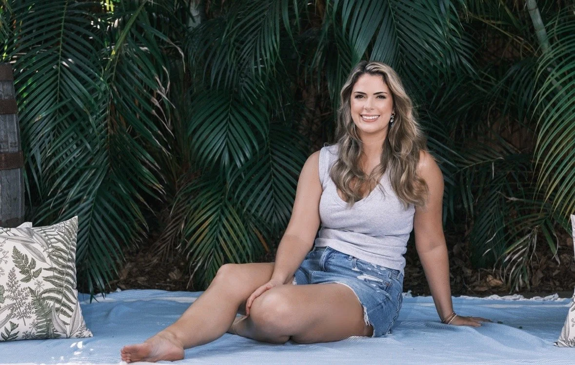 A woman sitting on a blanket outdoors surrounded by green foliage, smiling while looking to the side.