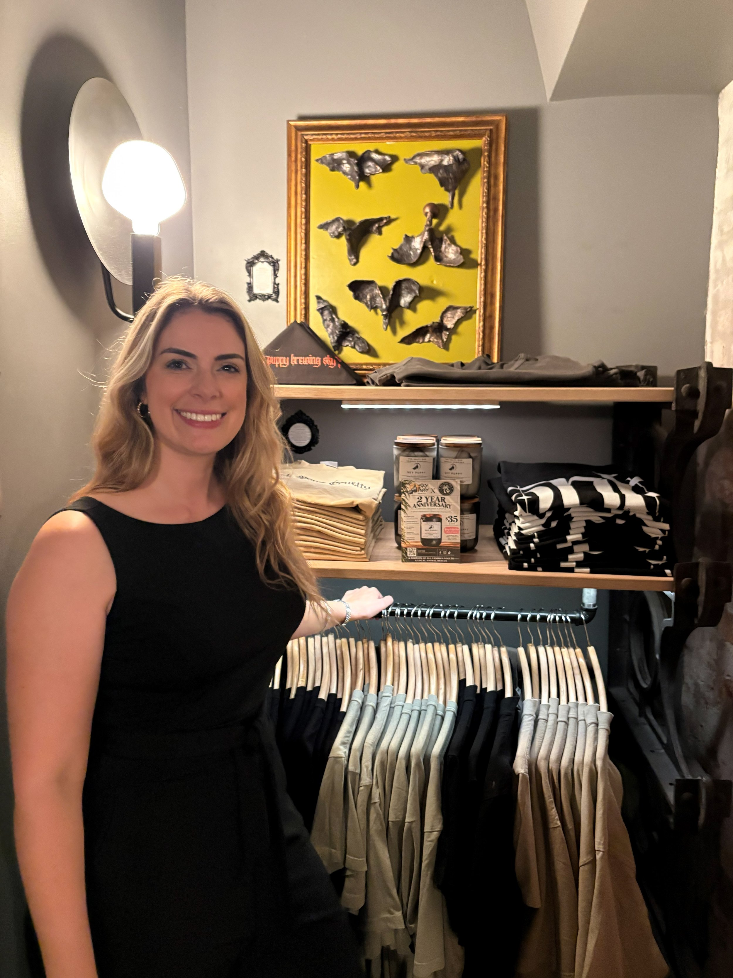 A woman with long blonde hair wearing a black sleeveless dress standing inside a clothing store near a rack with beige, gray, and black clothing. Behind her is a shelf with folded clothing, candles, and framed artwork, and a yellow-green wall with a display of decorative metallic bats in various positions.