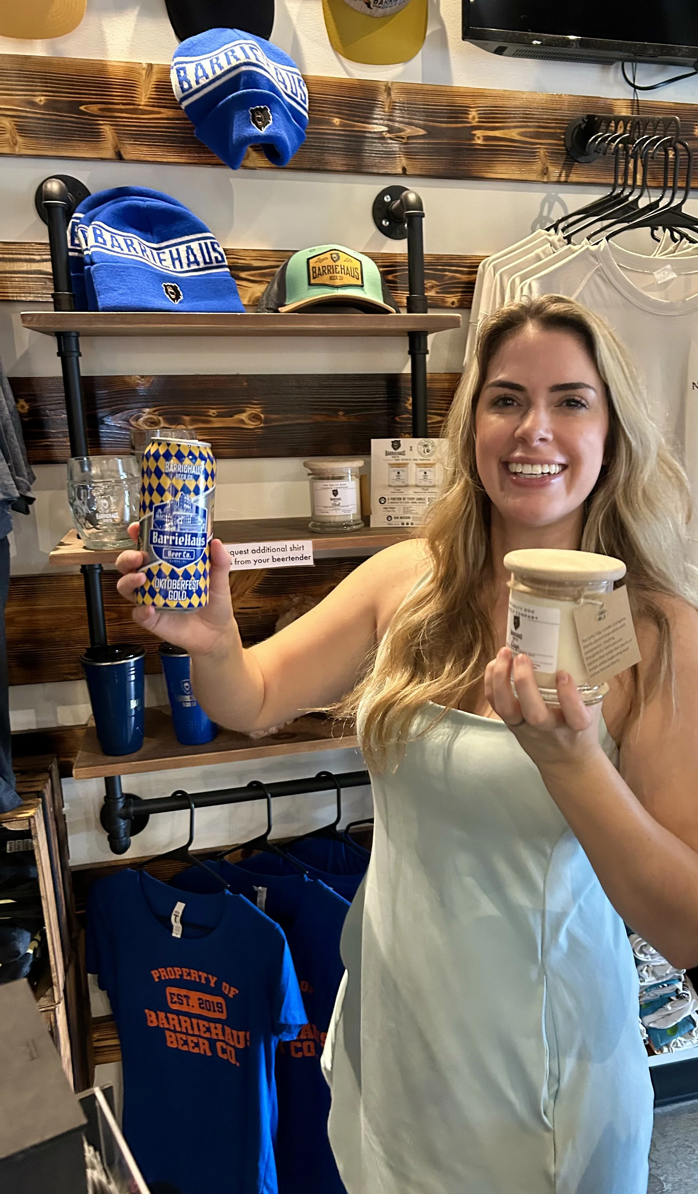 A woman with long blonde hair smiling and holding a beer can in a store. Behind her, shelves display blue and gray hats and a gray cap, white T-shirts, blue T-shirts, and merchandise with the Barriehaus Beer Co. logo.