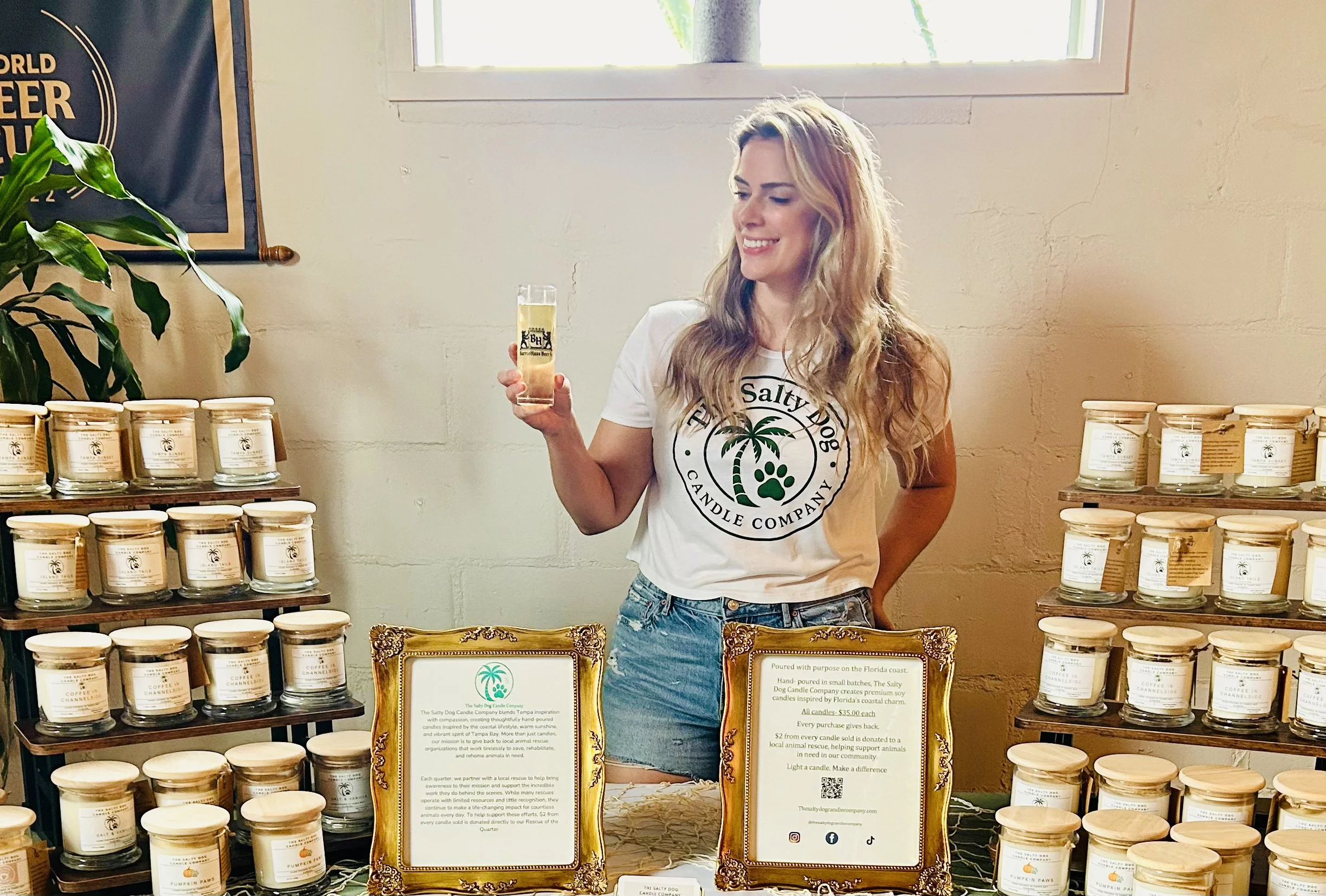A woman with long blonde hair smiling and holding a glass of beer at a booth displaying candles in jars. The booth has informational signs and several shelves filled with candles labeled with scents. The woman is wearing a white T-shirt with a palm tree and text logo, and denim shorts. There is a window and a wall with a poster in the background.