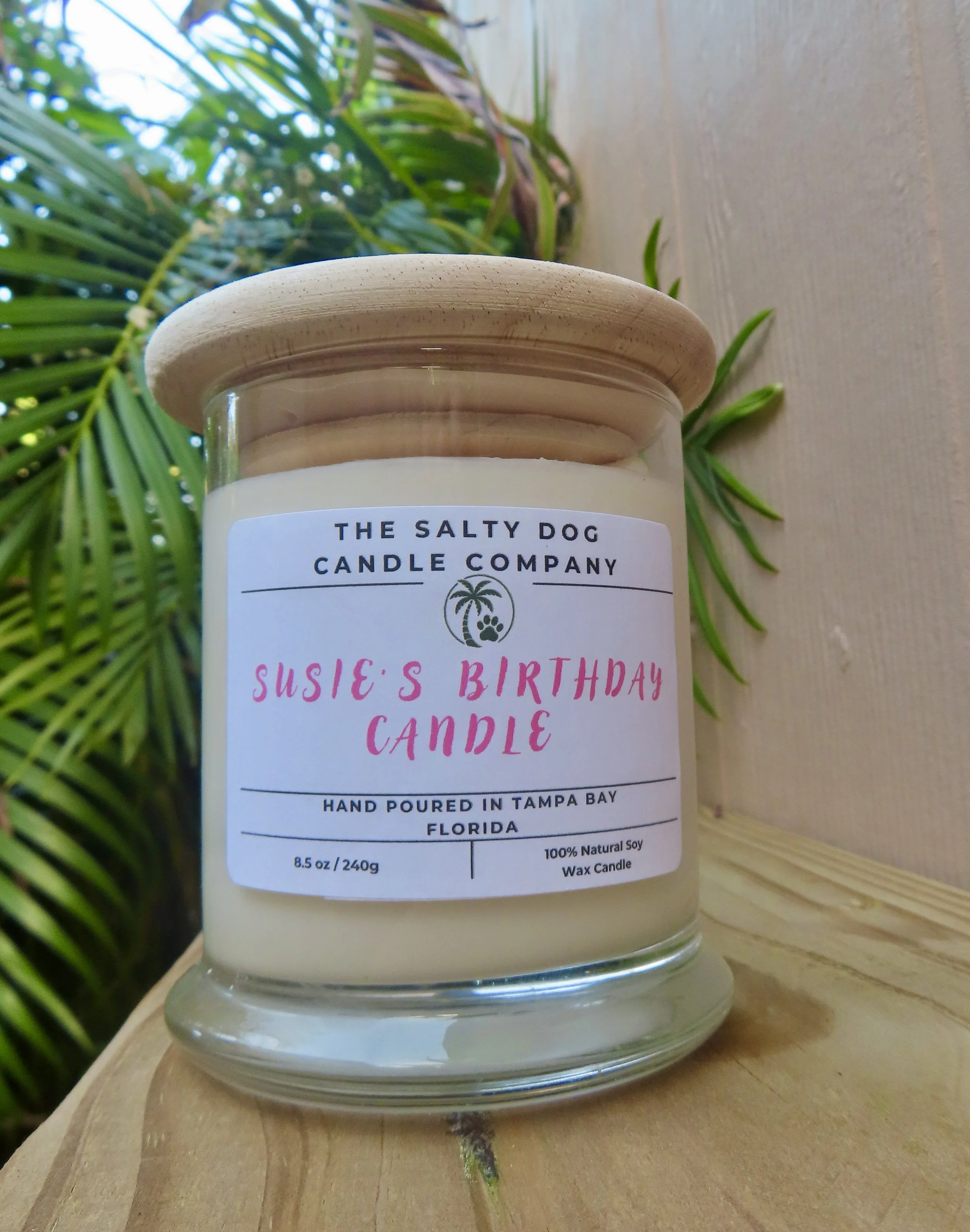 A candle labeled "Susie's Birthday Candles" produced by The Salty Dog Candle Company, placed on a wooden surface with a leafy green plant in the background.