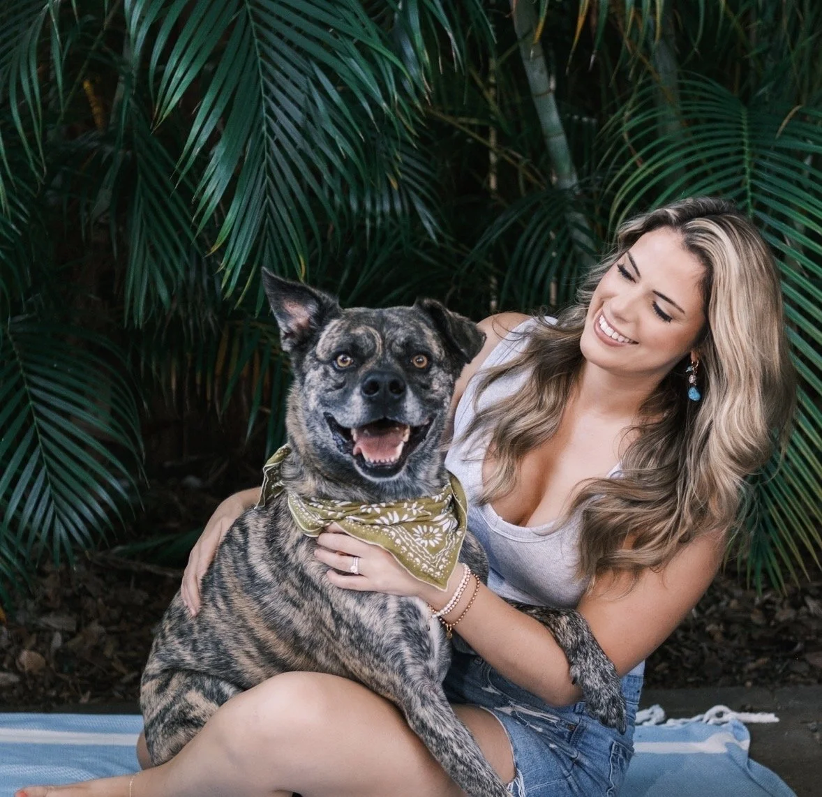 A smiling woman sitting on a blanket holding a happy brindle dog wearing a bandana, surrounded by lush green tropical plants.