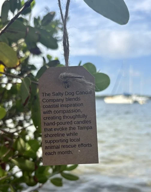 A close-up of a hanging tag from The Salty Dog Candle Company, with a beach and sailboats visible in the background. The tag describes the company's coastal-inspired candles that support animal rescue efforts.