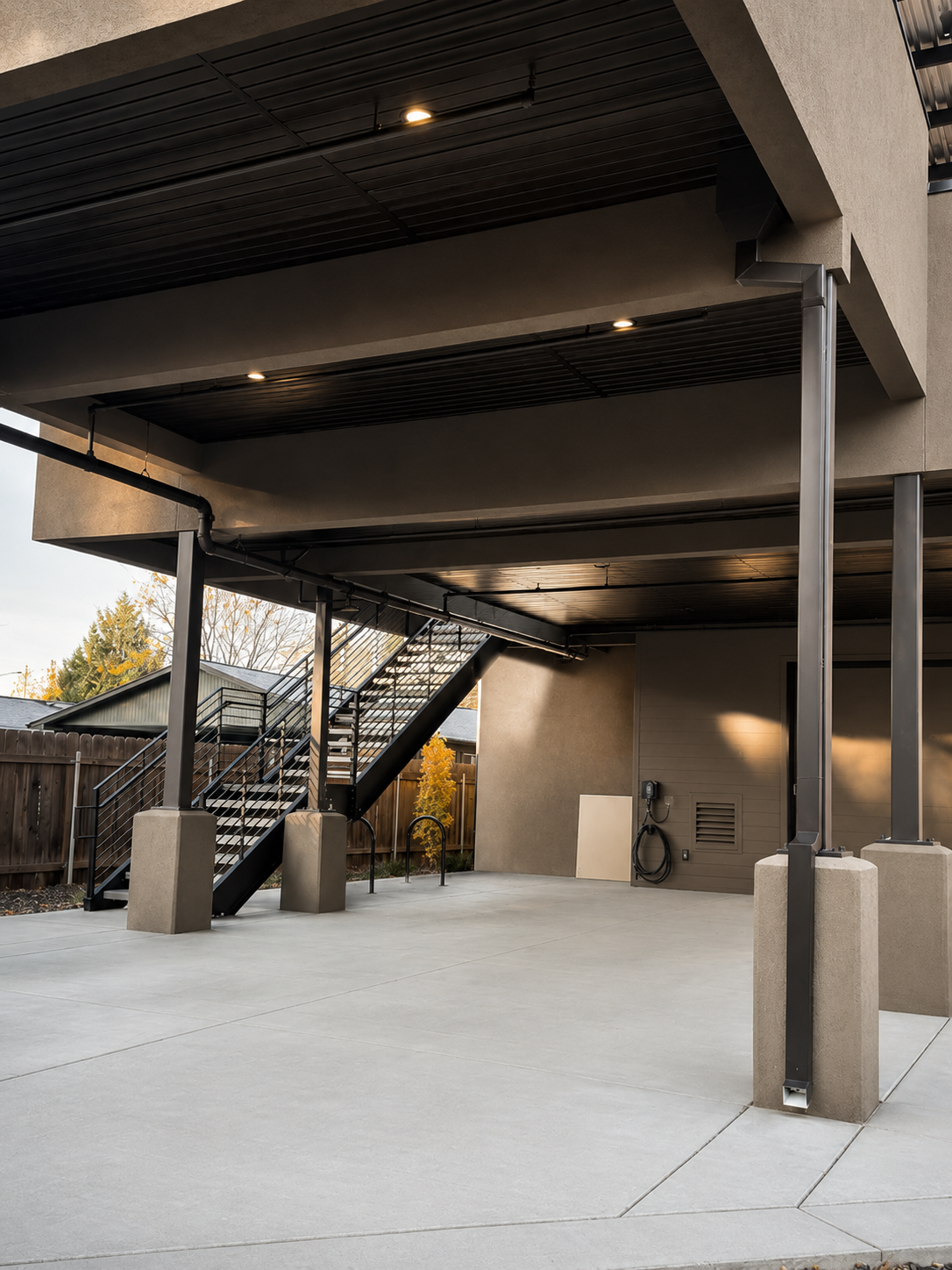 A modern apartment building exterior with a staircase leading to the upper level, concrete flooring, and a fenced backyard with trees.