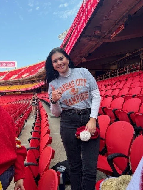 A young woman standing in a stadium filled with red seats, smiling at the camera, wearing a gray 'Kansas City Champions' sweatshirt, giving a thumbs-up, with a purse and a drink nearby.