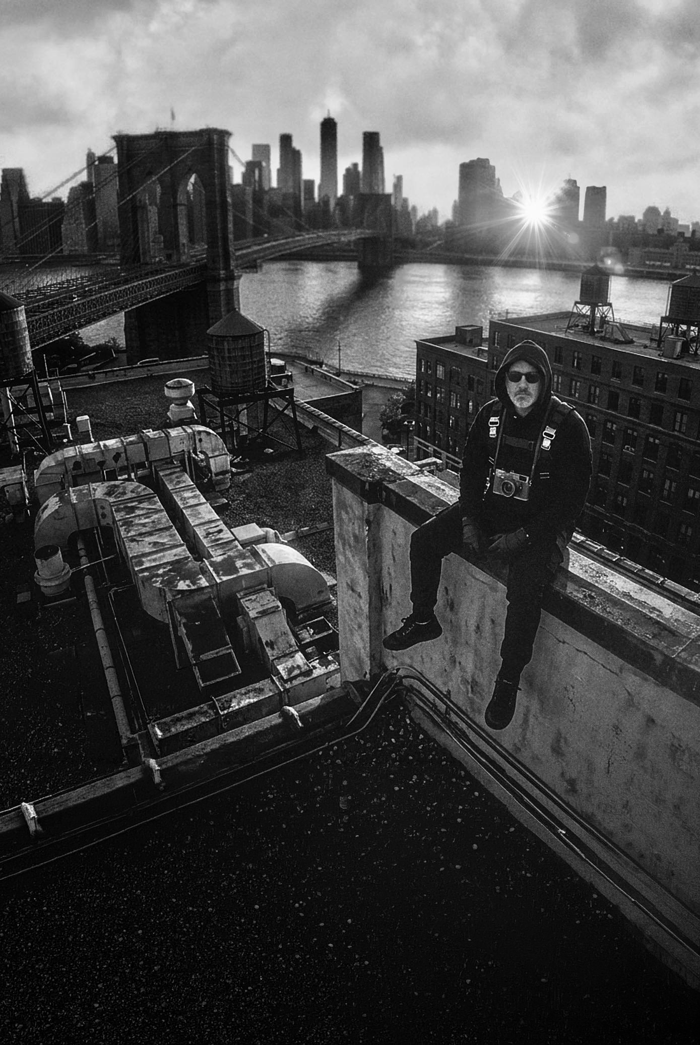 A man wearing sunglasses and a hoodie sitting on the edge of a rooftop in New York City, with the Brooklyn Bridge and Manhattan skyline in the background, black and white photo.