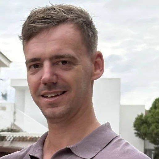 A man with short, light brown hair smiling outdoors, wearing a gray polo shirt, with a light-colored building and trees in the background.