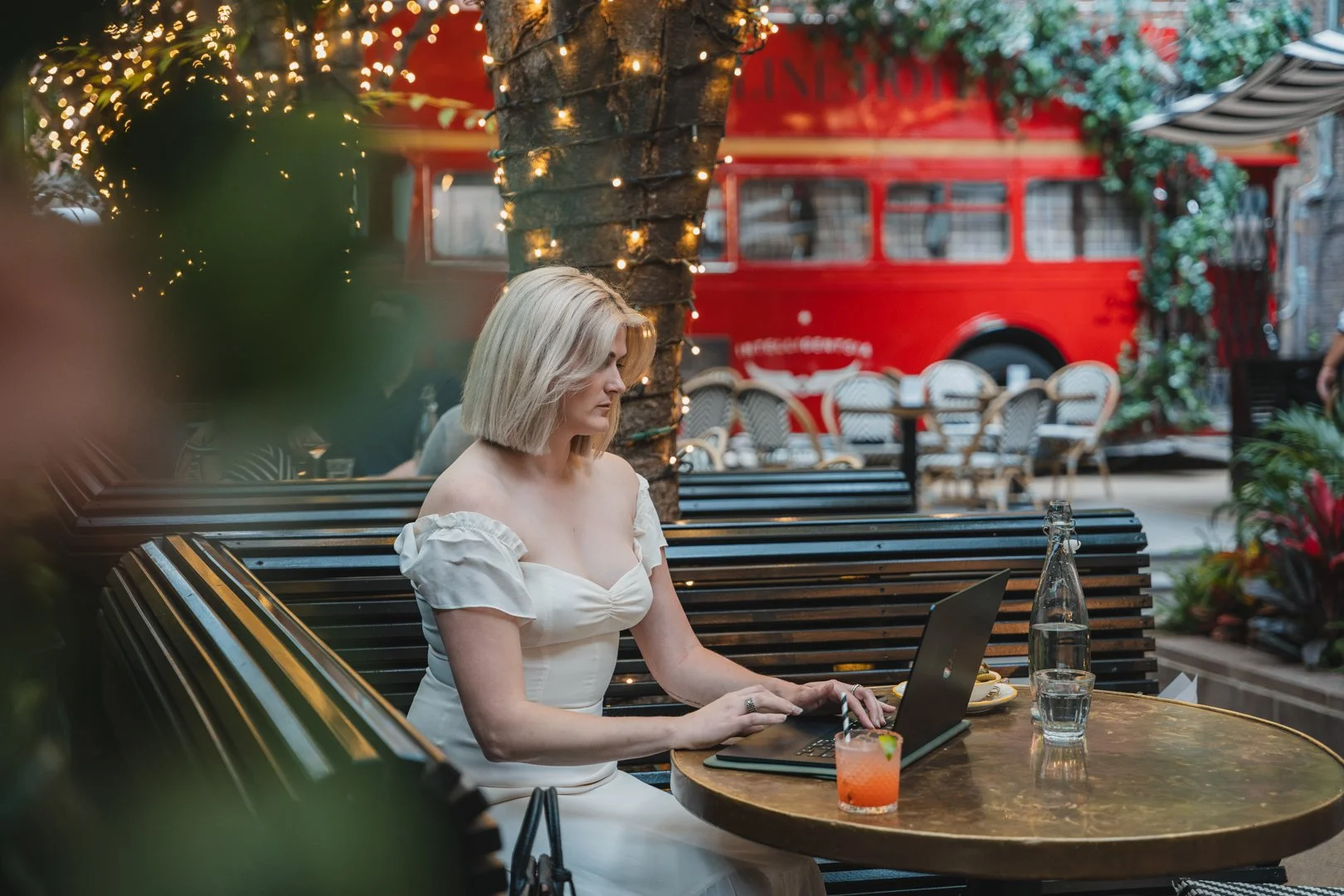 A woman with blond hair in a white off-shoulder dress sitting outside at a round table with a glass of water, a bottle of water, a small drink with a lime, and a laptop. She is working on her laptop. Behind her, there are benches, chairs, a tree wrapped with string lights, and a red double-decker bus in the background.