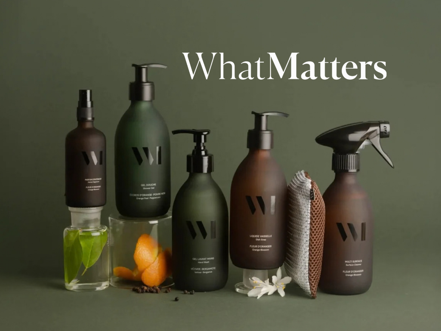 Collection of five dark bottles with pump and spray dispensers labeled with black text, accompanied by a transparent glass with leaves, a glass with kumquats and peppercorns, a striped cloth, and a white flower against an olive-green background, with