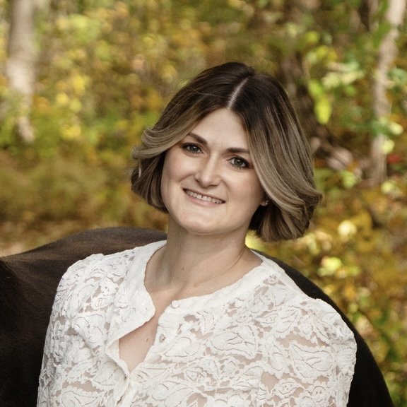 A woman with short, wavy brown hair smiling outdoors with autumn-colored trees in the background.
