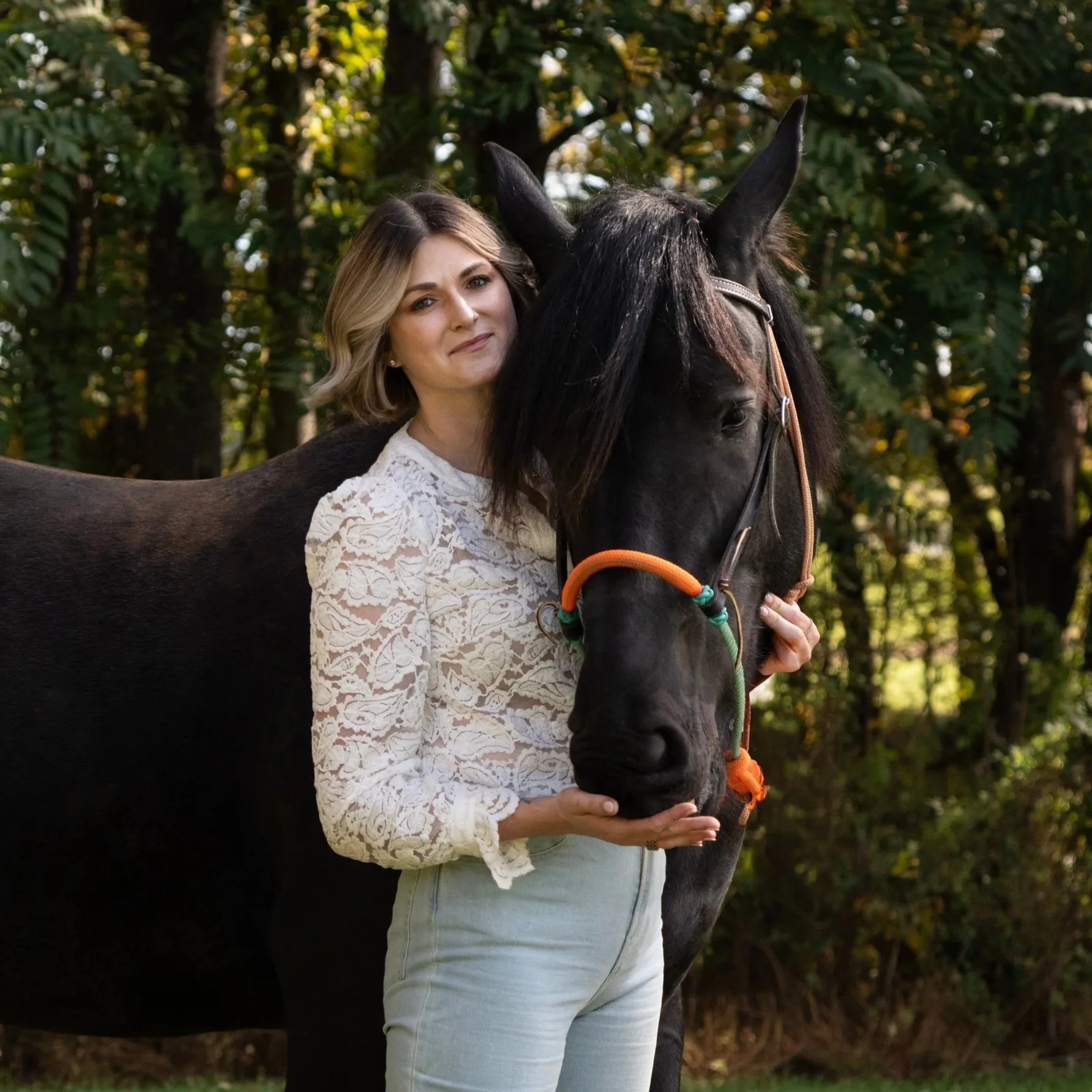 Woman wearing a white lace top, holding a black horse with an orange bridle in a wooded area with trees and sunlight.