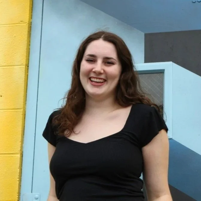 A young woman with long brown hair wears a black top, smiling outdoors in front of a colorful mural with blue, yellow, and gray sections.