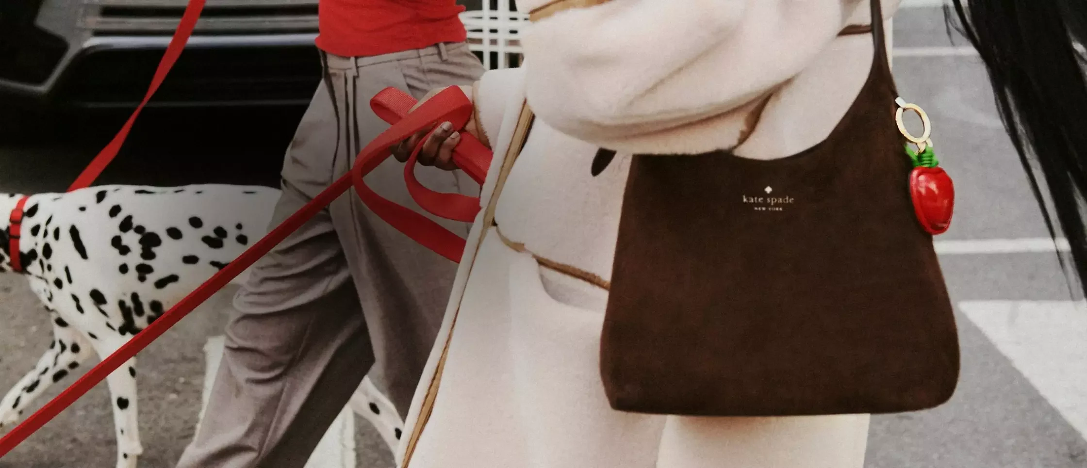 Close-up of a person holding a Kate Spade brown handbag with a chili pepper-shaped keychain, with another person in gray pants and a Dalmatian dog nearby.