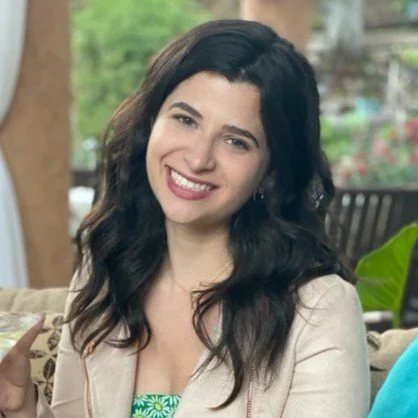 A woman with long dark hair smiling outdoors, sitting on a bench surrounded by greenery.