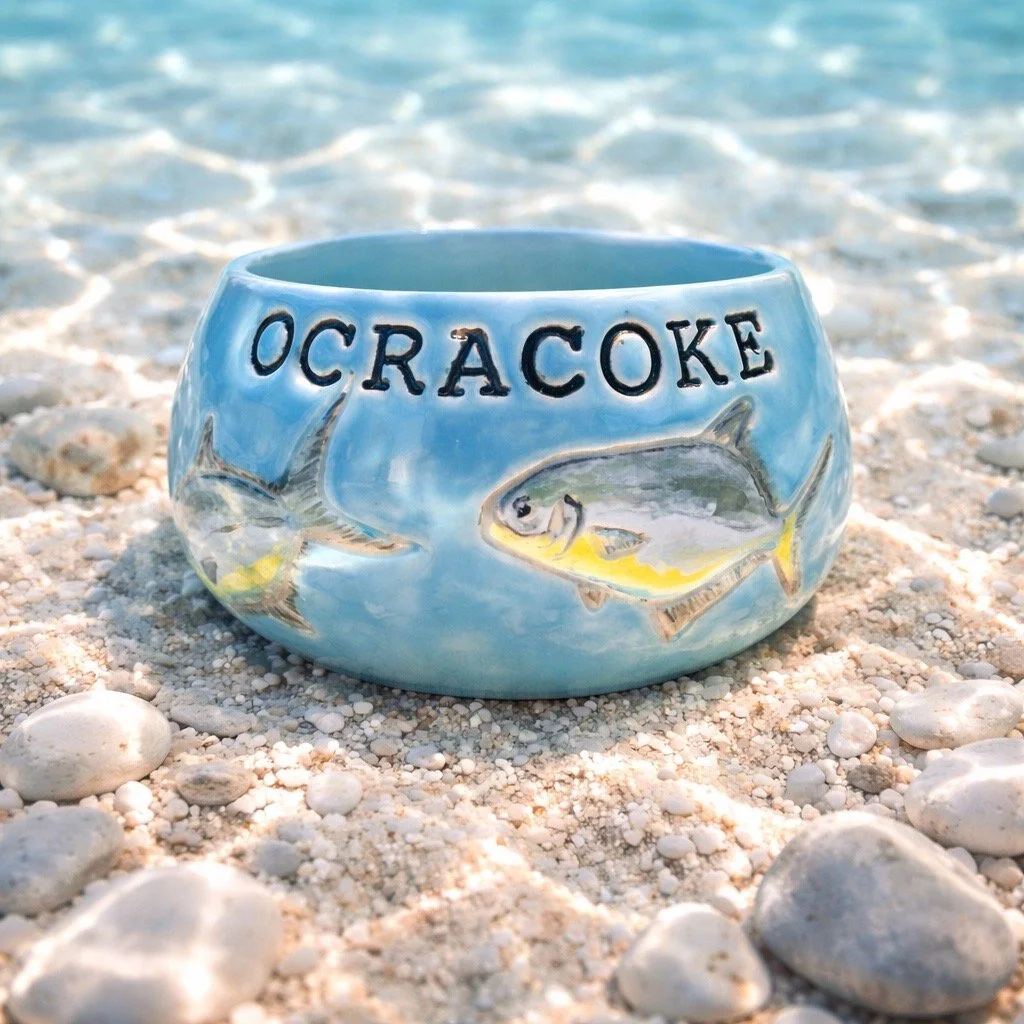 A ceramic fishbowl with the word 'Ocracoke' and painted fish illustrations, placed on a sandy beach near the water.