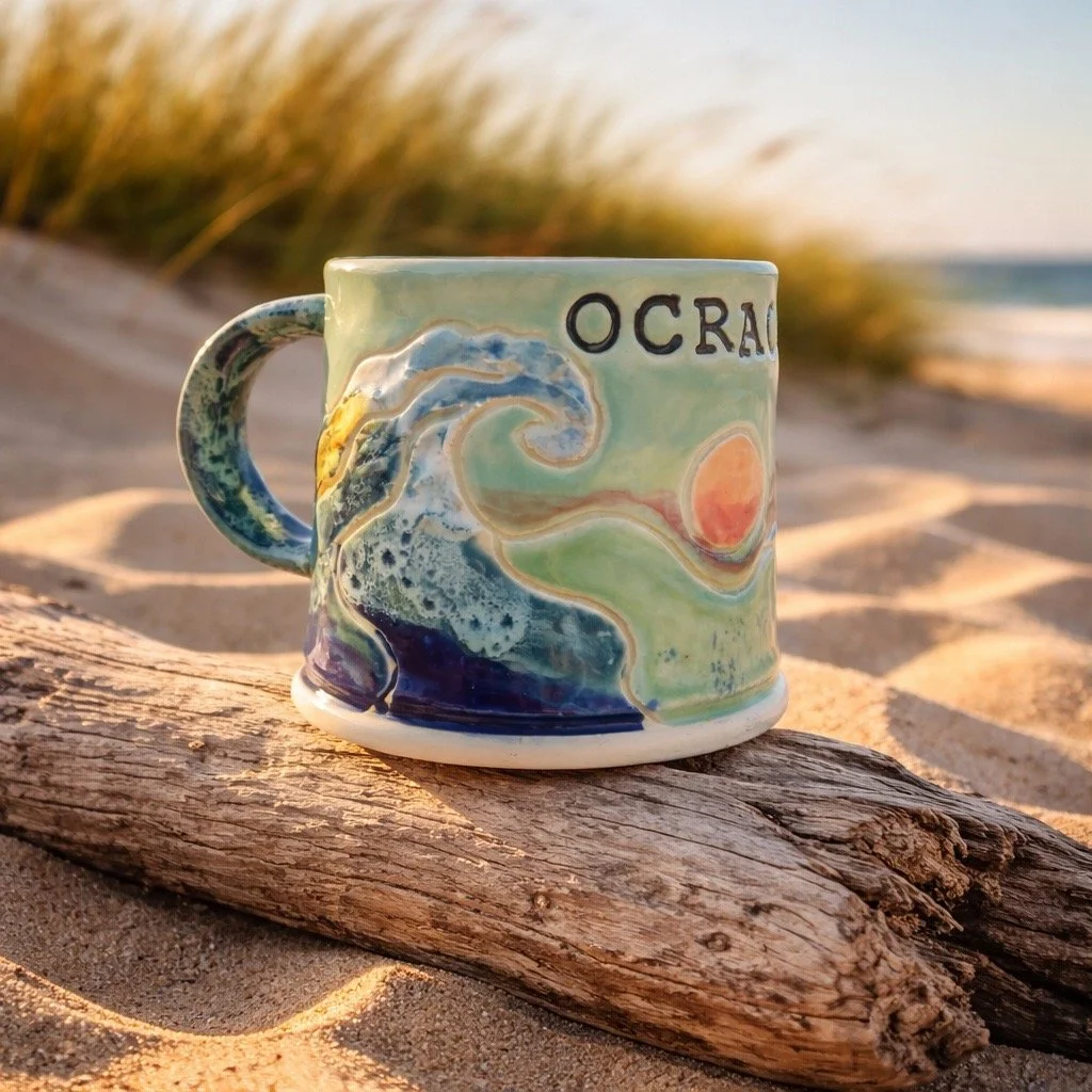 Colorful ceramic mug with ocean wave design and the partially visible word "OCEAN" on a log on a sandy beach, with grass and the ocean in the background.