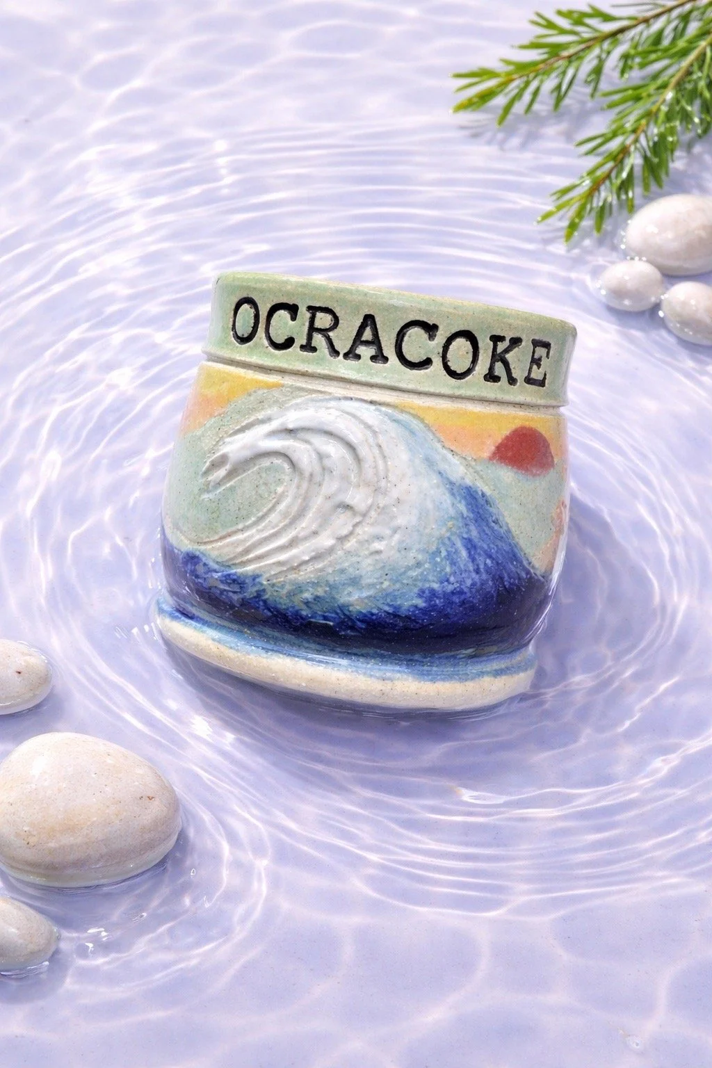 Decorative ceramic piece with a wave design and the word OCRACOKE on top, floating in water with white stones and a green pine branch nearby.