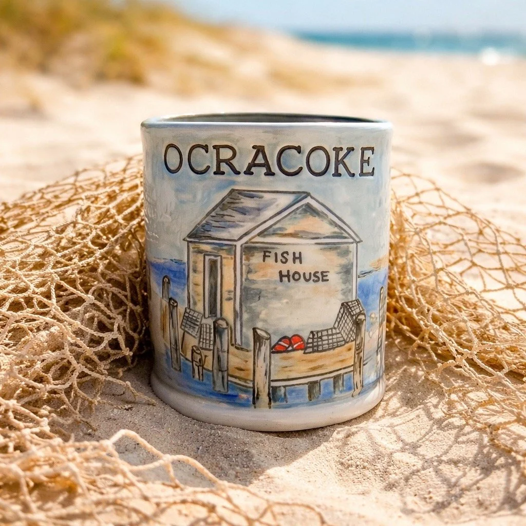 Ceramic mug with a beach scene and the words "Ocracoke Fish House," placed on the sand with a fishing net nearby, at the beach.