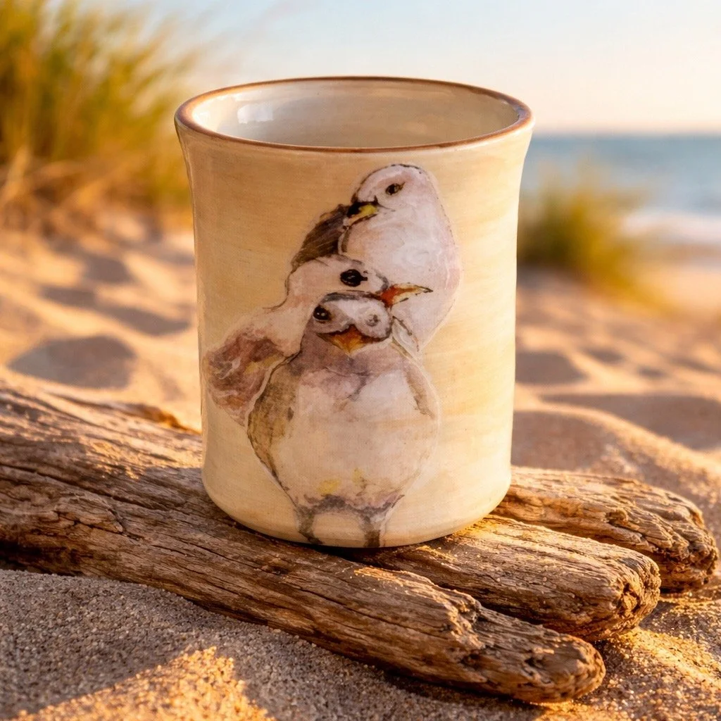 Ceramic mug with a painting of three birds, placed on a piece of driftwood on sandy beach, with blurred grass and ocean in background.