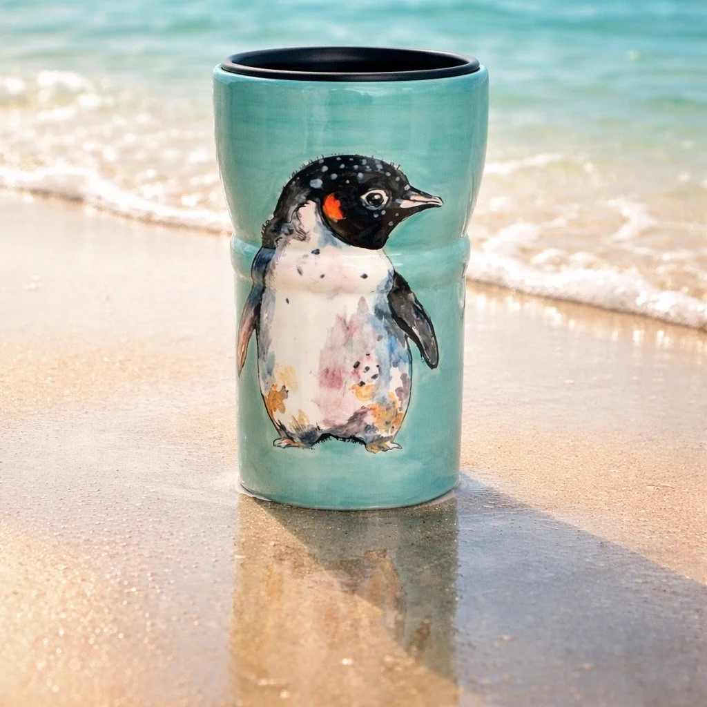Hand-painted ceramic mug with a cute penguin illustration, placed on sandy beach near the ocean.