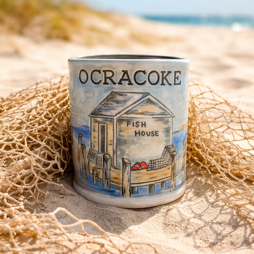 A ceramic mug with a coastal scene of a fish house, labeled 'Ocracoke,' on a sandy beach with seashells and fishing nets, with the ocean and sky in the background.