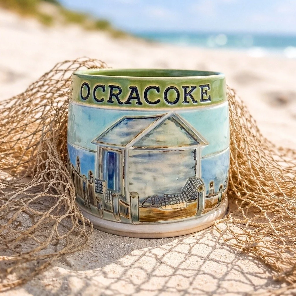 A ceramic mug with the word 'Ocracoke' and a painted scene of a beach with a dock, umbrellas, and the ocean, placed on the sand next to a fishing net at the beach.