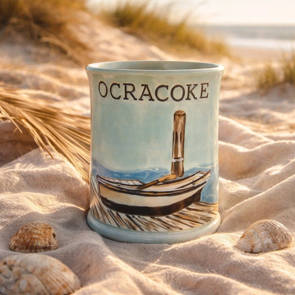 A mug with the word 'OCRACOKE' written on it, featuring a painted picture of a boat on the beach. The mug is placed on sand with seashells, a dried palm leaf, and blurred background of a beach scene.