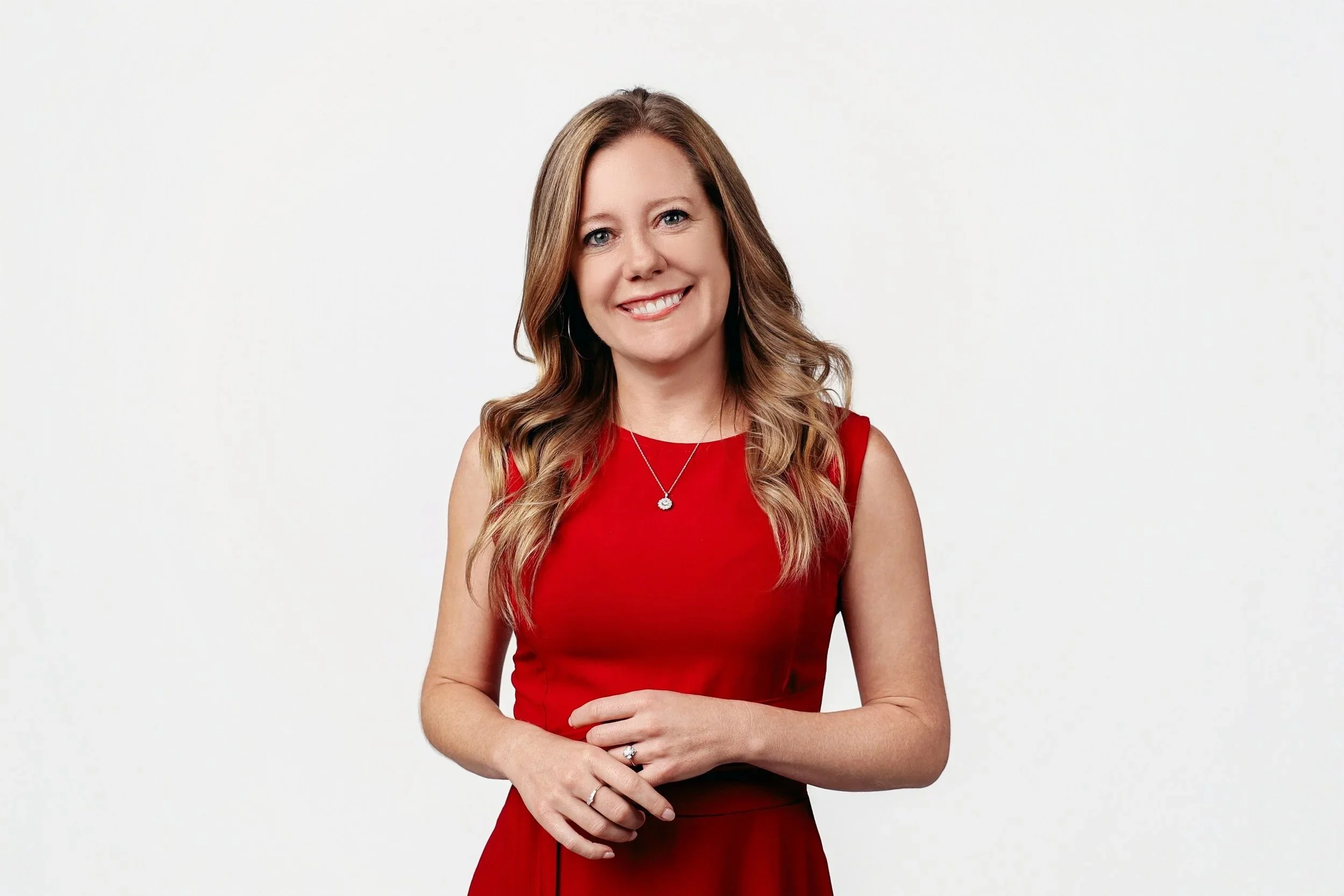 A woman with long, wavy brown hair, wearing a red sleeveless dress, standing against a white background, smiling at the camera.